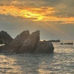Three rocks jut out of a calm sea, against a deep orange sunset. Popular destination for October half term getaways.