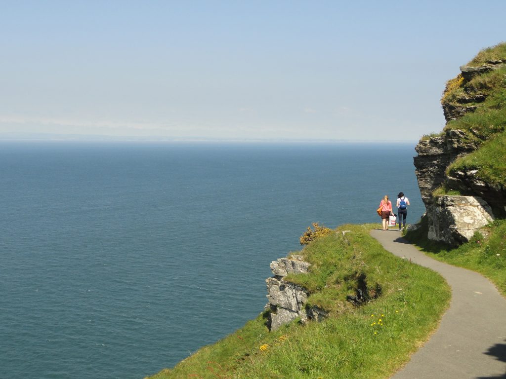 A footpath curved around a sheer cliff face, looks out over a blue sea and sky.